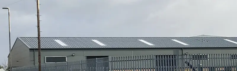 Industrial unit with a grey profiled metal roof and evenly spaced rooflights, behind palisade fencing